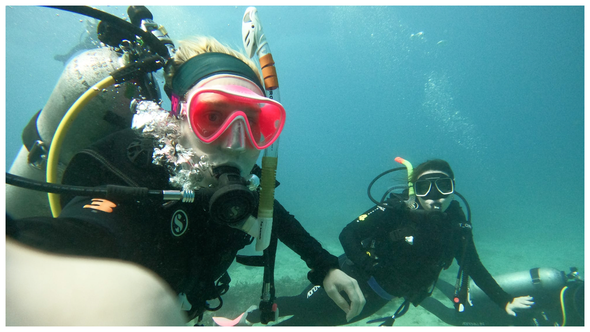 Two divers underwater; one takes a selfie with a pink mask, while the other poses nearby, both wearing black wetsuits.
